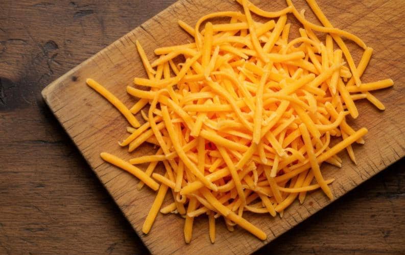 Ilustrasi keju cheddar berwarna kuning-oranye dengan beberapa potongan keju di atas latar belakang putih.