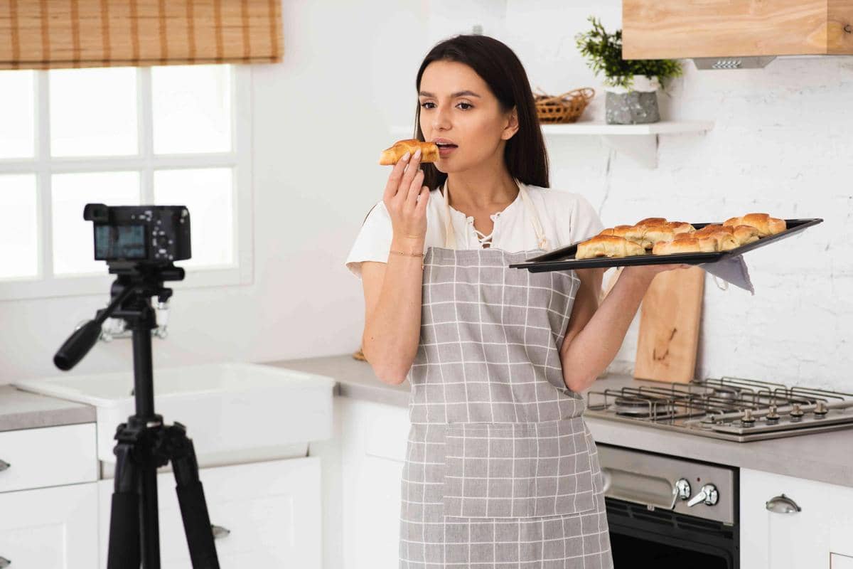 Seorang perempuan muda merekam proses memasak di dapur untuk membuat konten produk menggunakan ponsel di tripod.