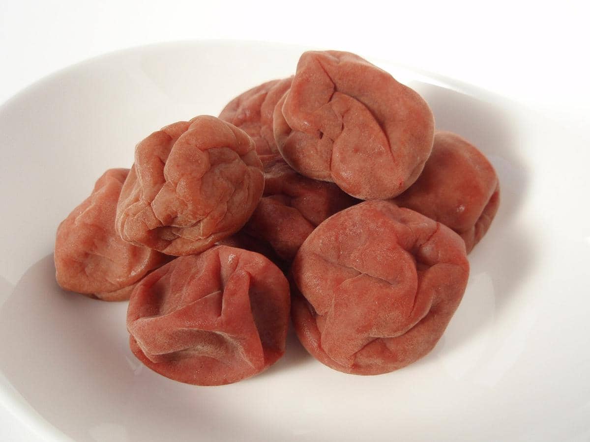 Ilustrasi umeboshi, acar plum Jepang berwarna merah kecokelatan, disajikan dalam mangkuk kecil di atas latar putih.