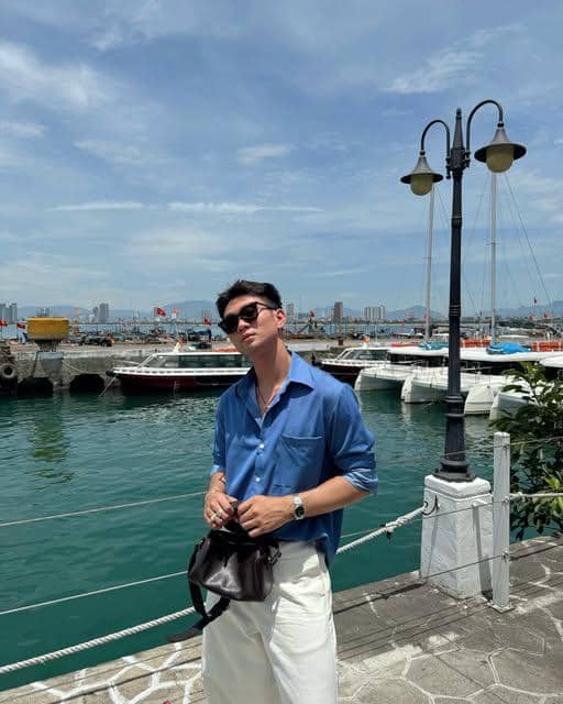 Vo Khanh Hung tampil dengan OOTD pantai sambil berpose di tepi laut, mengenakan pakaian kasual bernuansa santai.