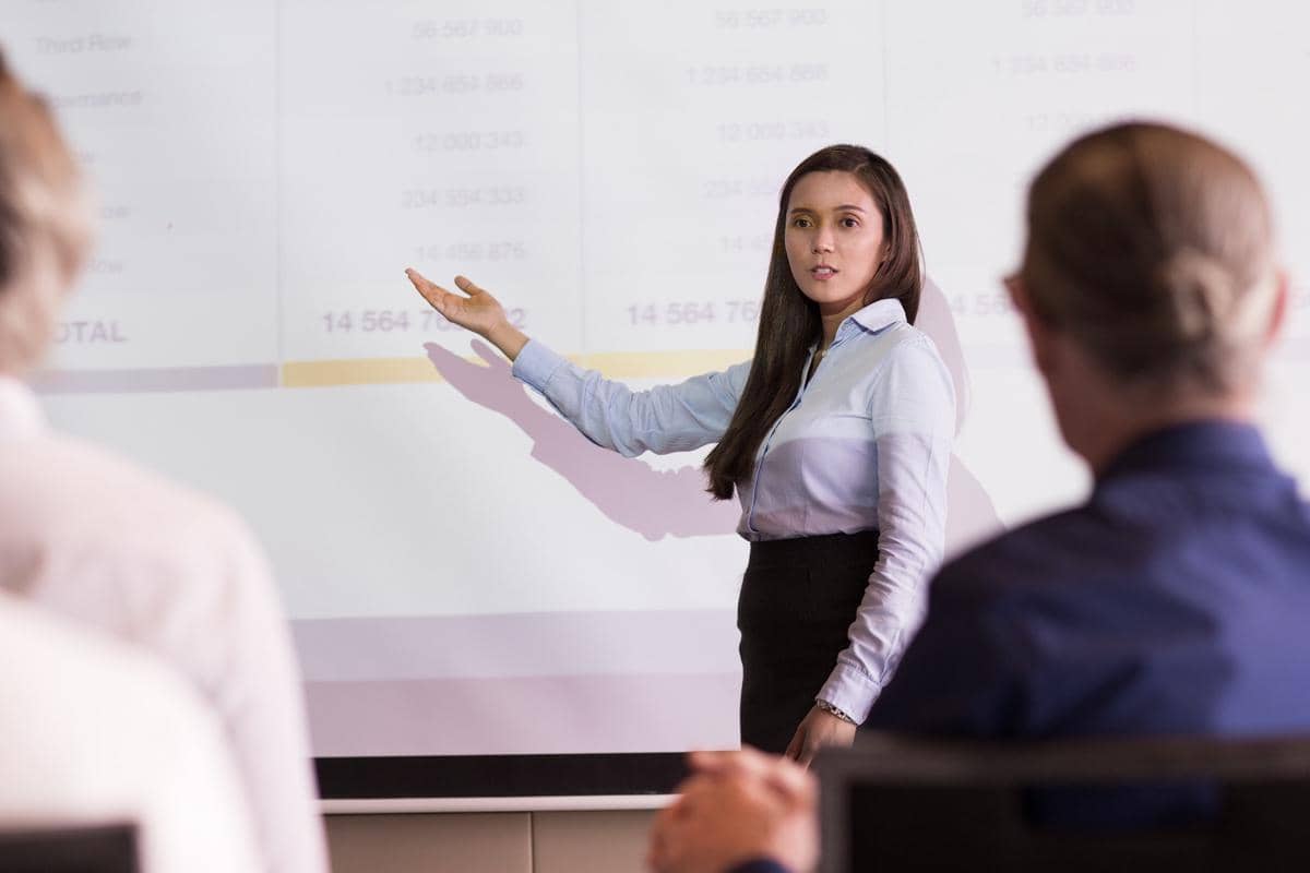 Seorang perempuan pebisnis berjas menjelaskan data di meja kepada audiens dalam ilustrasi presentasi bisnis.