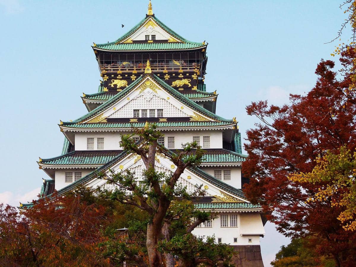 Panorama Osaka Castle Park di Osaka dengan bangunan kastel putih beratap hijau di antara pepohonan dan langit cerah.