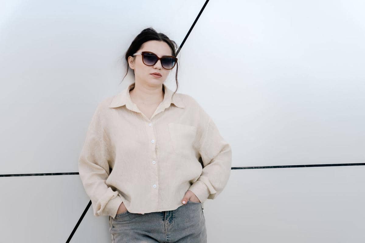 Seorang pria mengenakan kemeja oversized putih dan celana gelap, berdiri di dalam ruangan bernuansa modern.