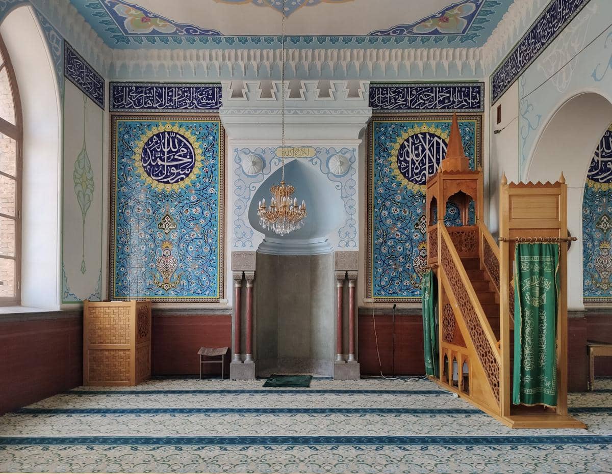 Interior aula salat Masjid Juma dengan mihrab, mimbar, serta lampu gantung yang menerangi ruang ibadah.
