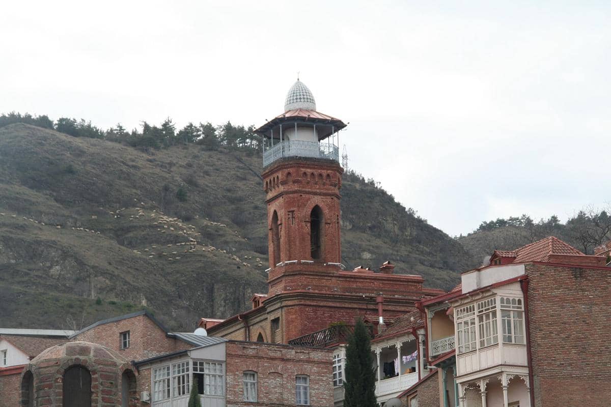 Menara Masjid Juma di Tbilisi menjulang dengan ornamen bata merah di antara bangunan dan pepohonan sekitar.