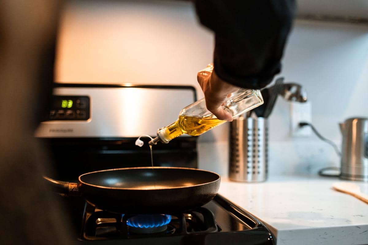 Tangan seseorang menuangkan minyak goreng dari botol ke wajan sebagai ilustrasi aktivitas memasak di dapur rumah.