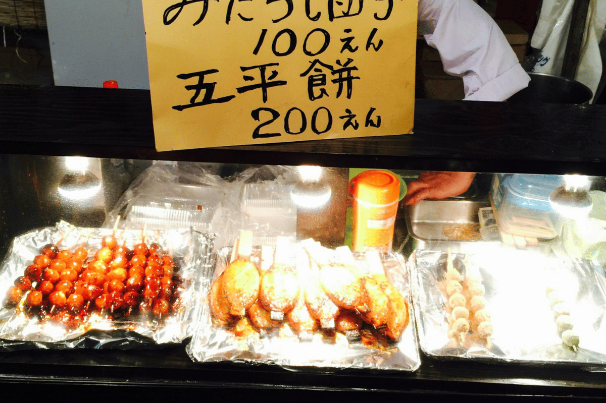 Dua tusuk gohei mochi berbentuk pipih dengan saus cokelat mengilap, disajikan di atas piring putih.