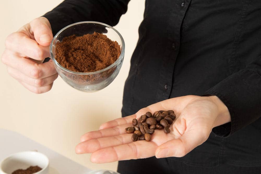 Ilustrasi bubuk kopi berwarna cokelat dalam mangkuk kayu, dikelilingi biji kopi dan daun hijau segar.