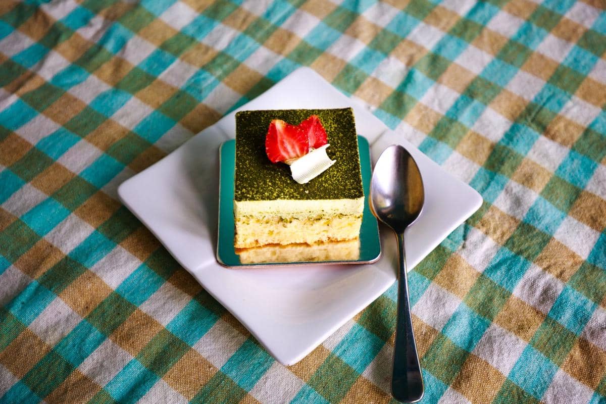 Segelas minuman ube matcha berlapis warna ungu dan hijau, disajikan dengan es di atas meja bernuansa hangat.