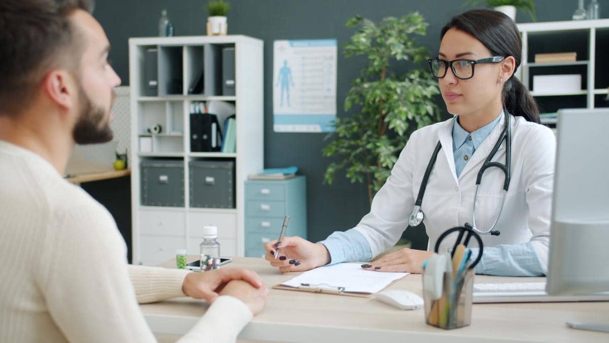Seorang pasien duduk berkonsultasi dengan dokter di ruang praktik, membahas kondisi kesehatan sambil melihat catatan medis.