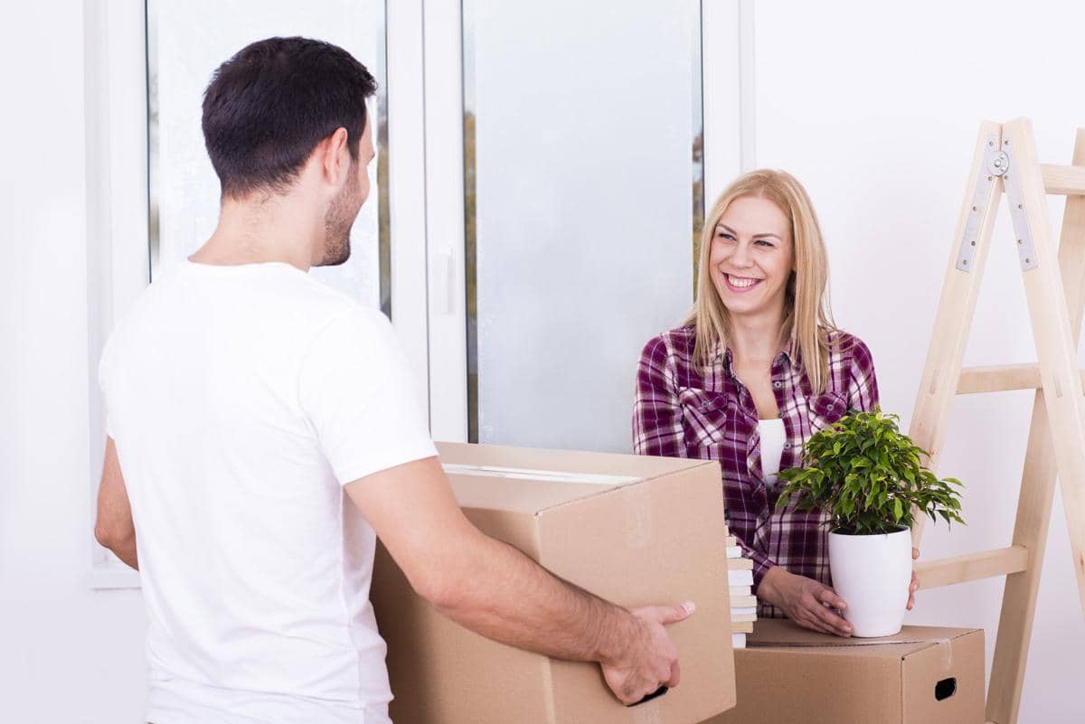 Pasangan sedang tersenyum membawa kardus saat pindah bersama ke rumah baru dengan latar ruangan yang masih kosong.