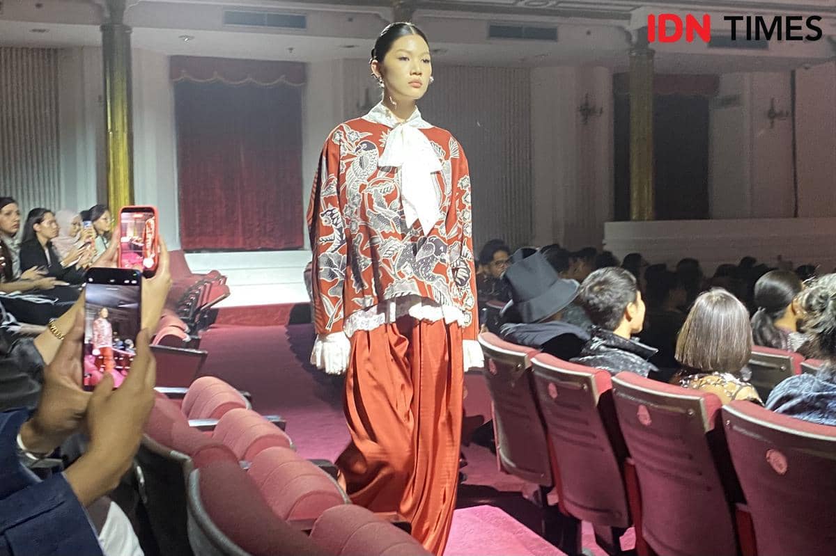 Atasan merah bermotif burung warna-warni dengan pita putih pada bagian depan sebagai detail busana yang manis.