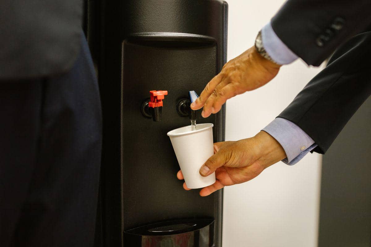 Dispenser air minum dengan keran untuk air dingin dan air panas, digunakan untuk mengisi gelas di ruangan.