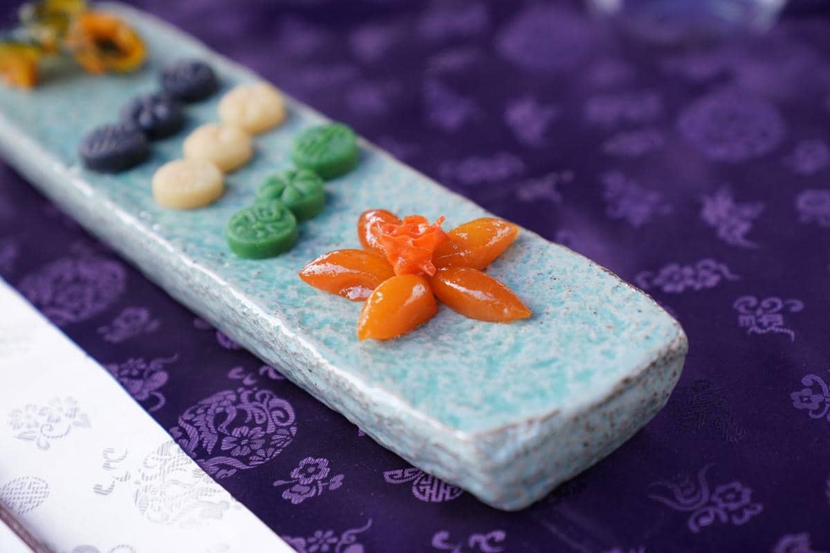 Ilustrasi songpyeon, kue beras Korea berbentuk setengah bulan berwarna pastel yang tersusun di atas piring.
