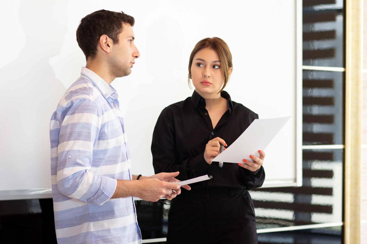 Seorang perempuan berdiri membaca dokumen saat dua rekan kerja duduk membahas grafik di meja dalam suasana presentasi kantor.