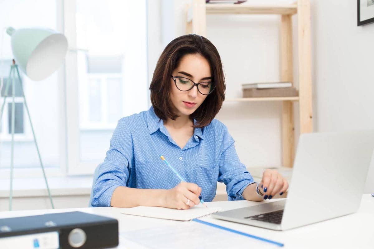 Perempuan berambut cokelat mengenakan kemeja biru dan kacamata menulis dengan serius di meja kerja kantor.