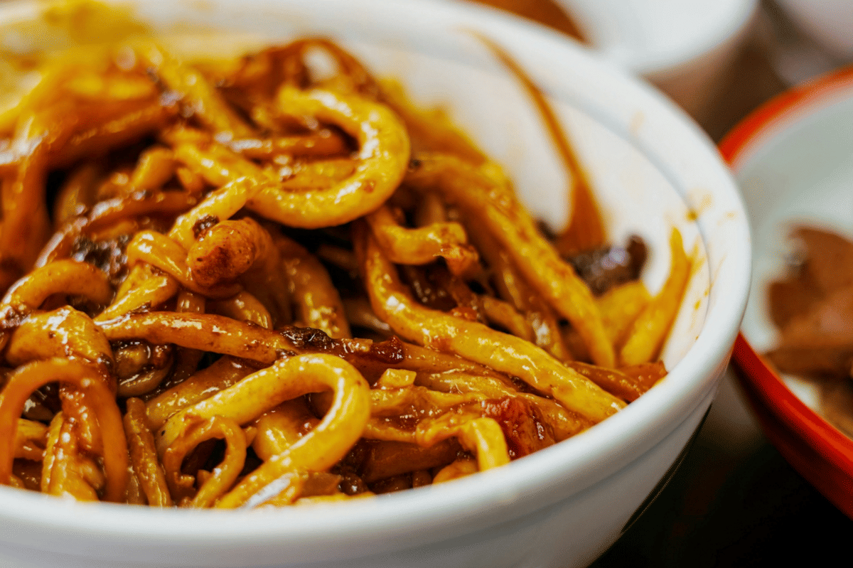 Semangkuk udon carbonara dengan chili oil, saus krim, dan taburan daun bawang dalam ilustrasi makanan.