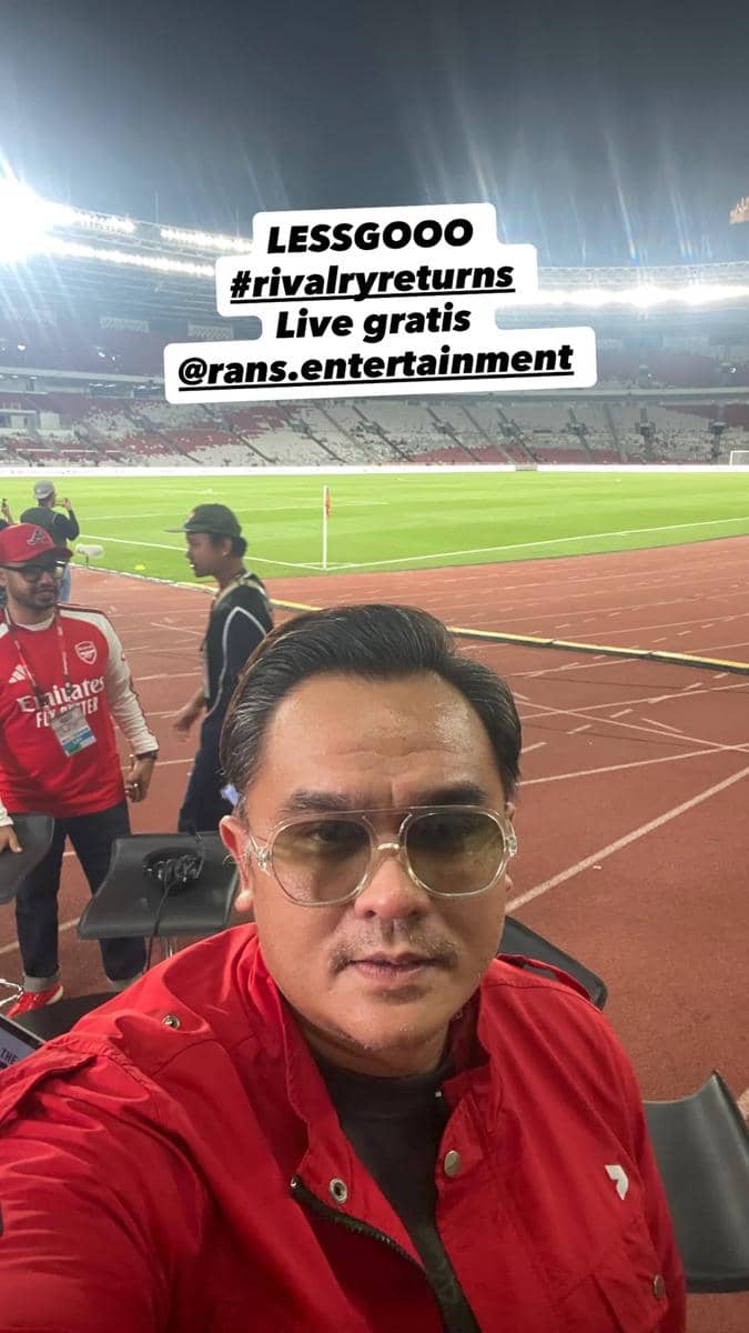 Seorang artis berpose bersama para pemain United Legends dan North London Legends di area stadion sepak bola.