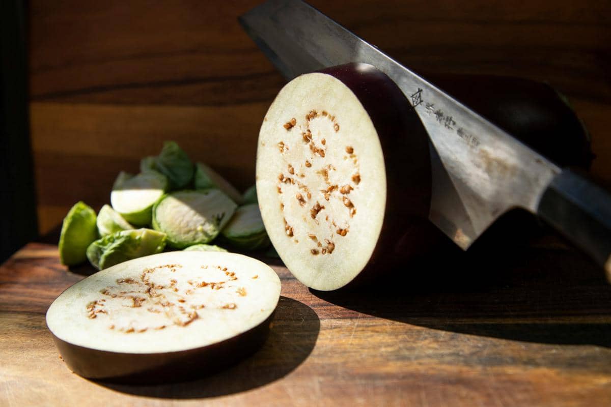 Seorang pria mengiris terong ungu di atas talenan kayu dengan pisau saat menyiapkan bahan masakan di dapur.