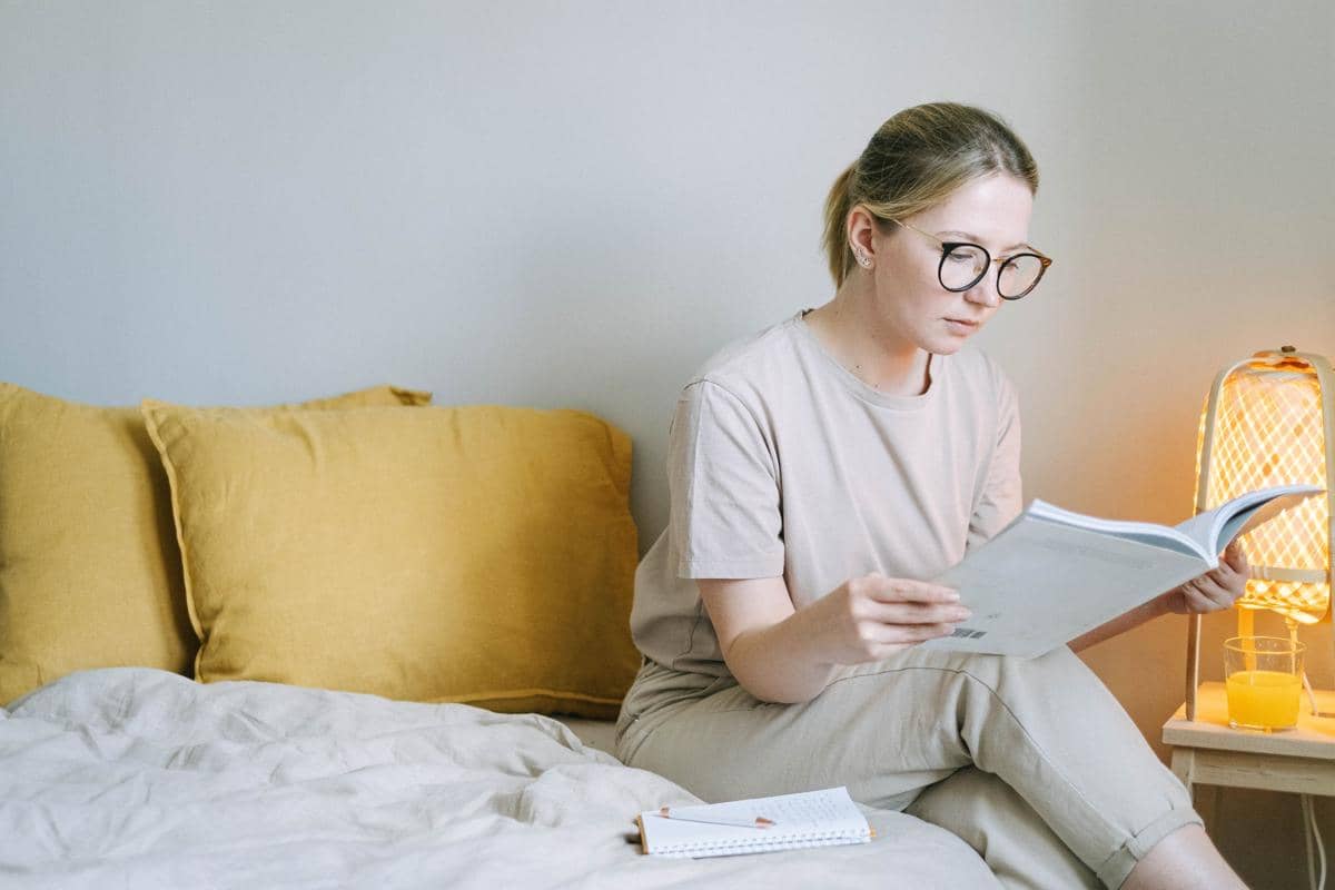 Ilustrasi seorang wanita bersantai membaca buku di kamar tidur untuk melepas stres secara sehat
