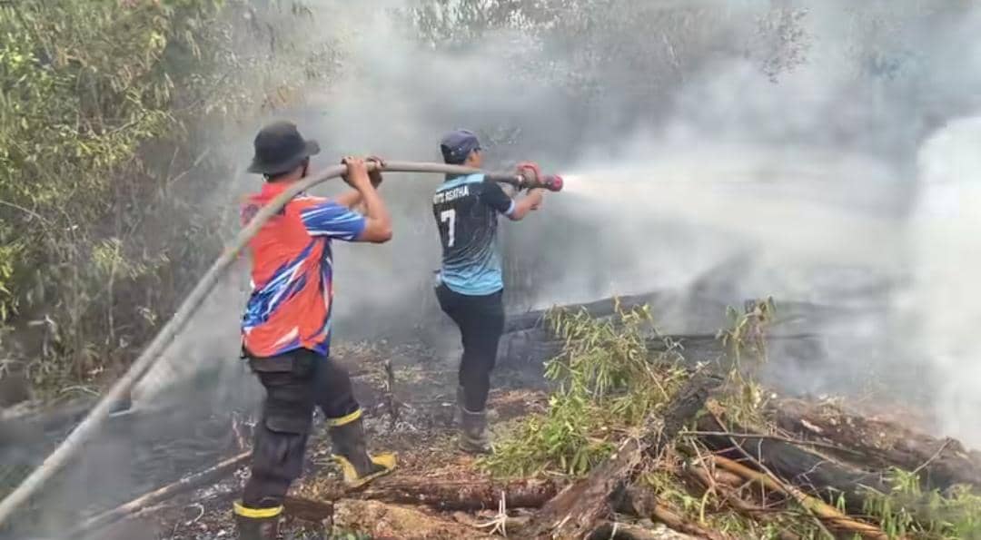 Petugas BPBD Muba memadamkan api di lahan terbakar menggunakan selang air, dengan asap mengepul di sekitar lokasi.