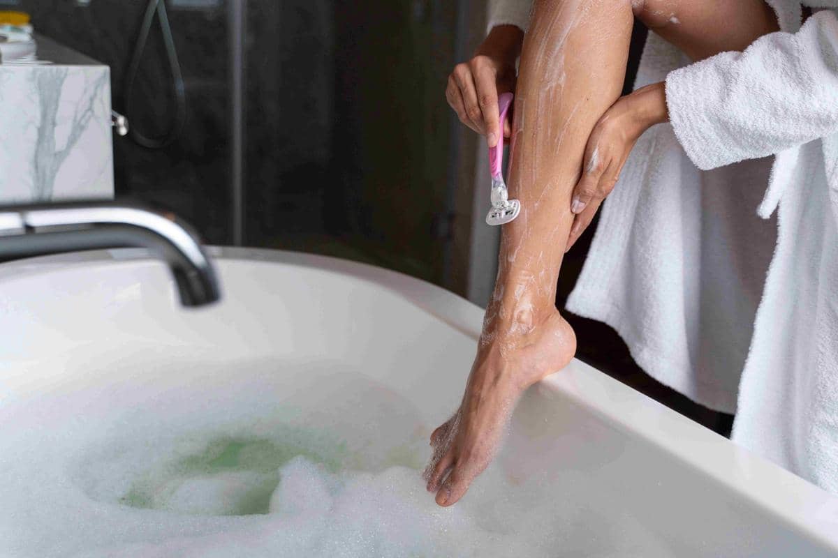 Seorang perempuan mencukur bulu kaki dengan alat cukur sebelum mandi sebagai ilustrasi membersihkan kaki.