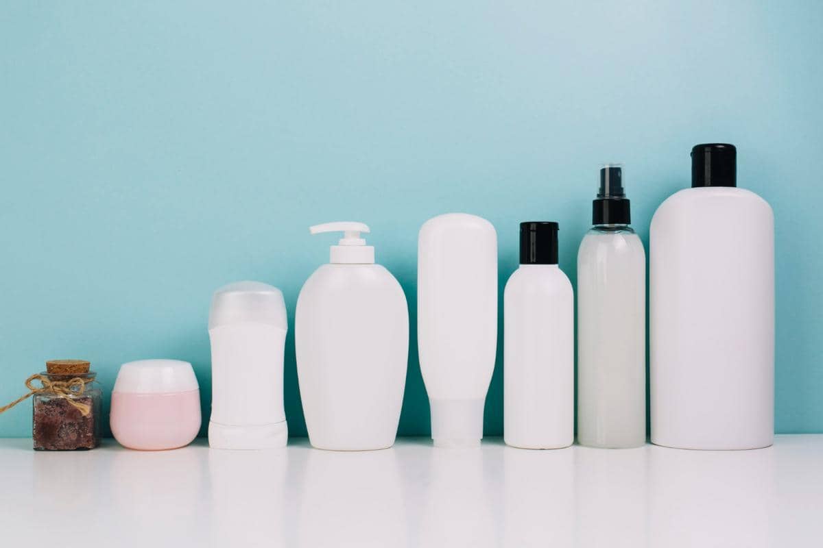 Deretan kemasan botol produk perawatan tubuh dan deodoran putih polos berlatar belakang biru pastel.