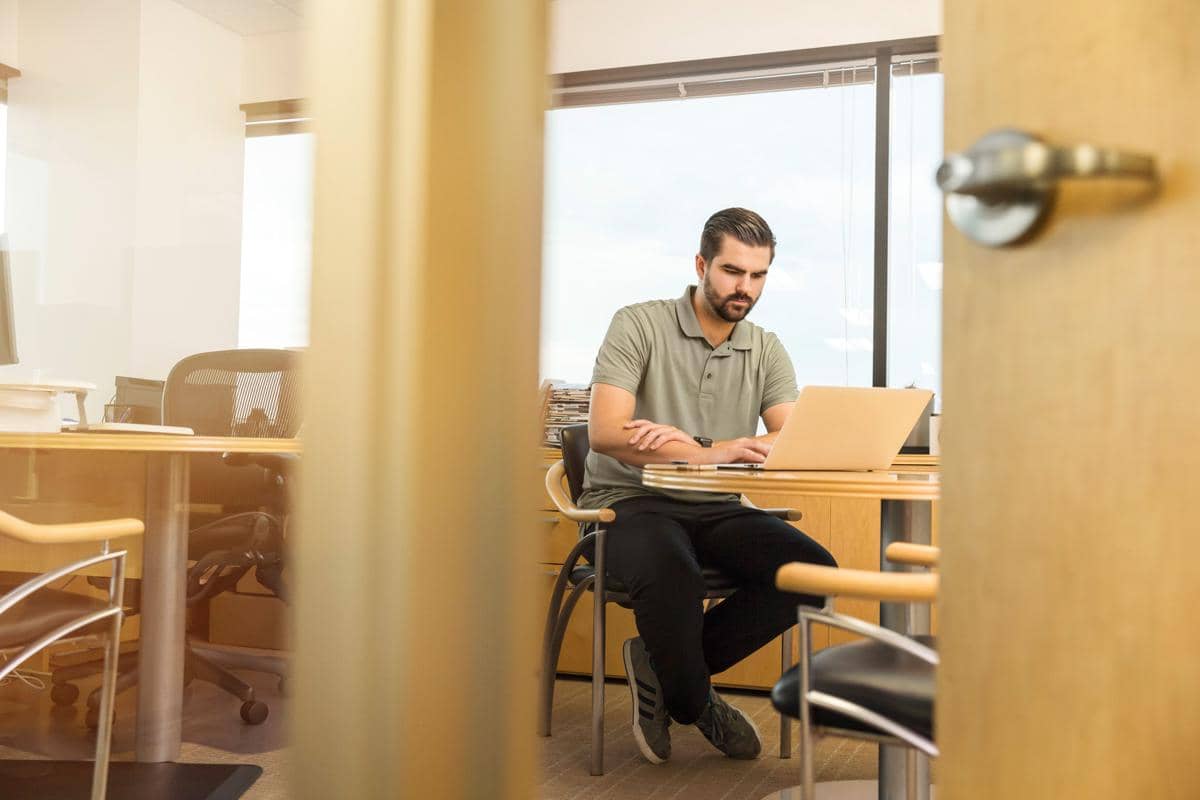 Seorang pria berkaus polo hijau duduk santai sambil bekerja menggunakan laptop di meja kantor.