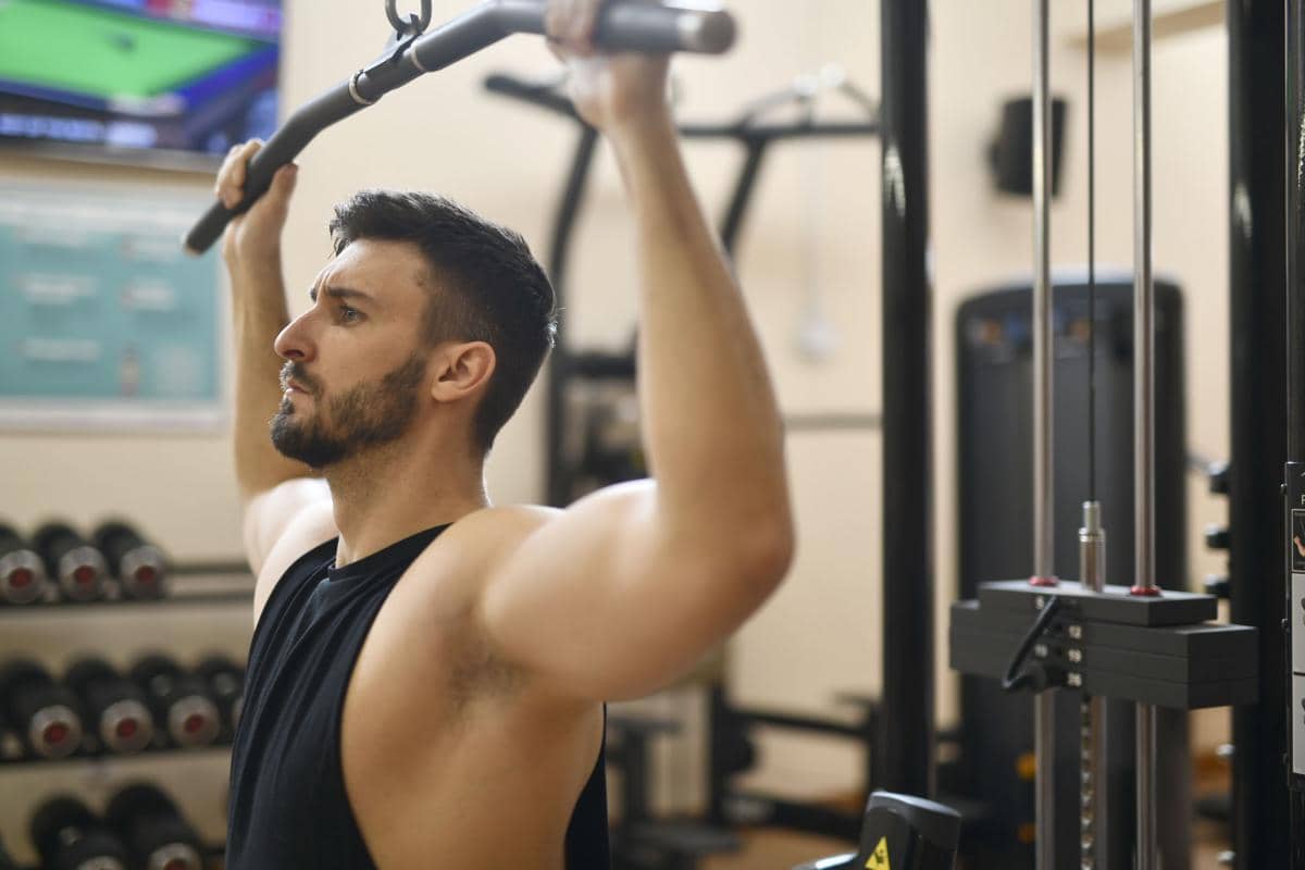 Seorang pria berotot mengenakan kaus tanpa lengan sedang menarik beban lat pulldown di gym.