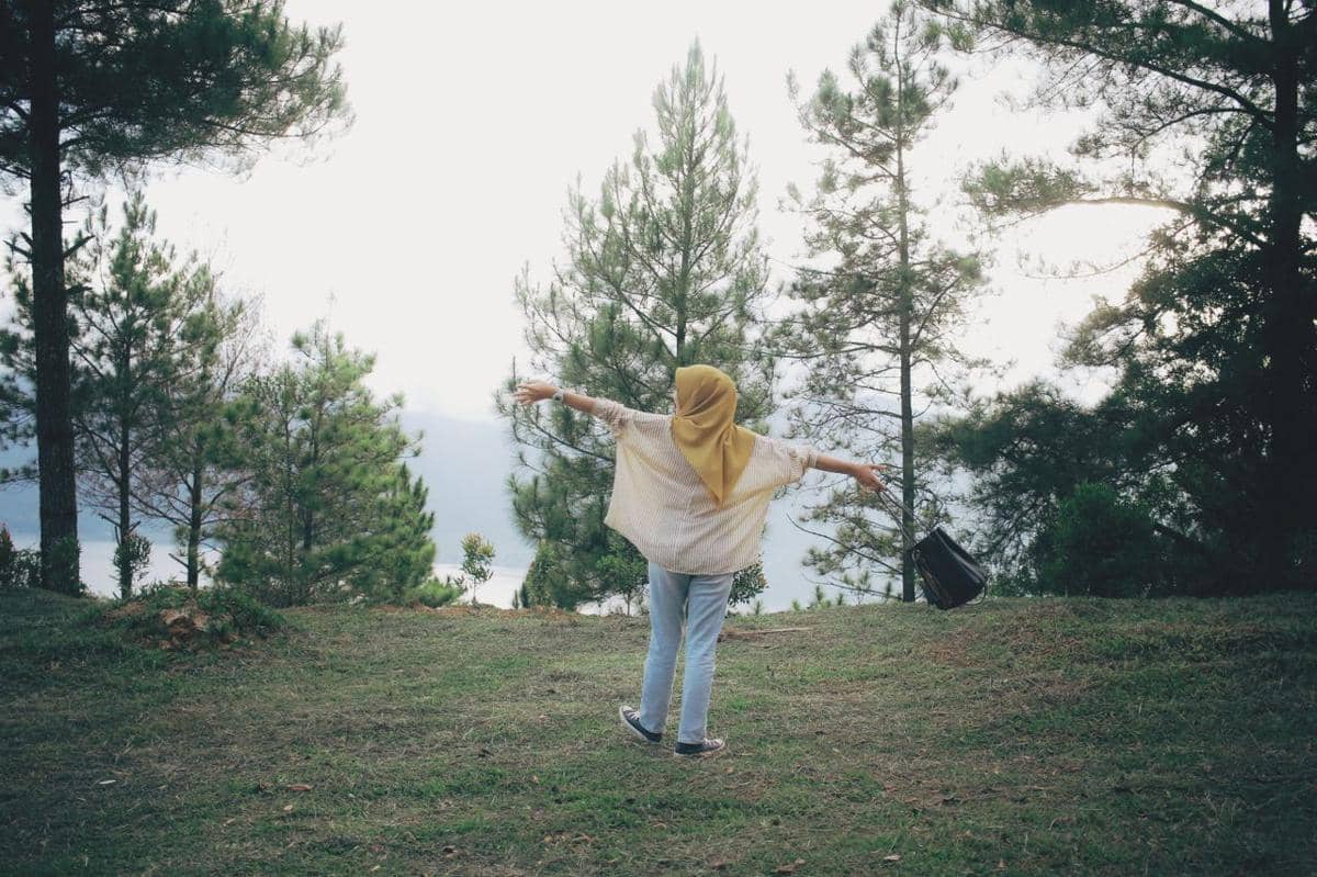 Seorang pria duduk di tepi danau memandang perbukitan hijau dan langit cerah, menikmati keindahan alam.