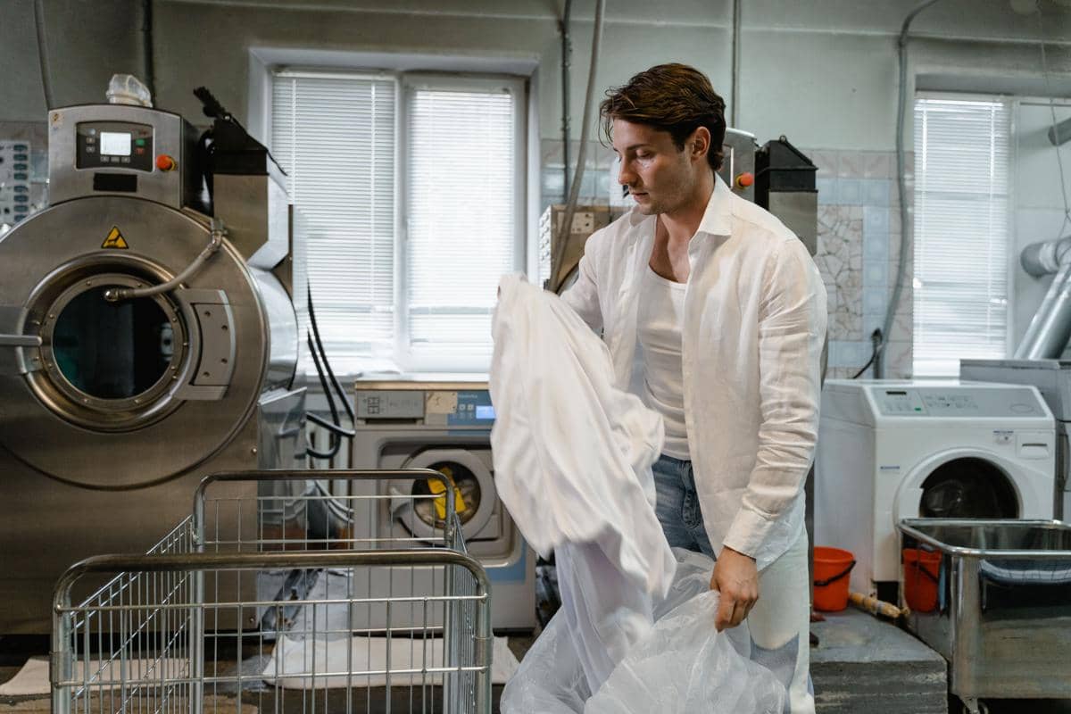 Seorang pria kemeja putih berdiri di ruang cuci laundry memegang pakaian putih di dekat mesin cuci industri.
