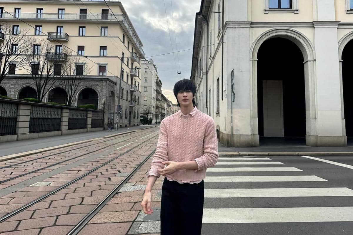 Byeon Woo Seok berjalan di penyeberangan jalan memakai cable knit sweater warna pink di atas kemeja dan celana hitam