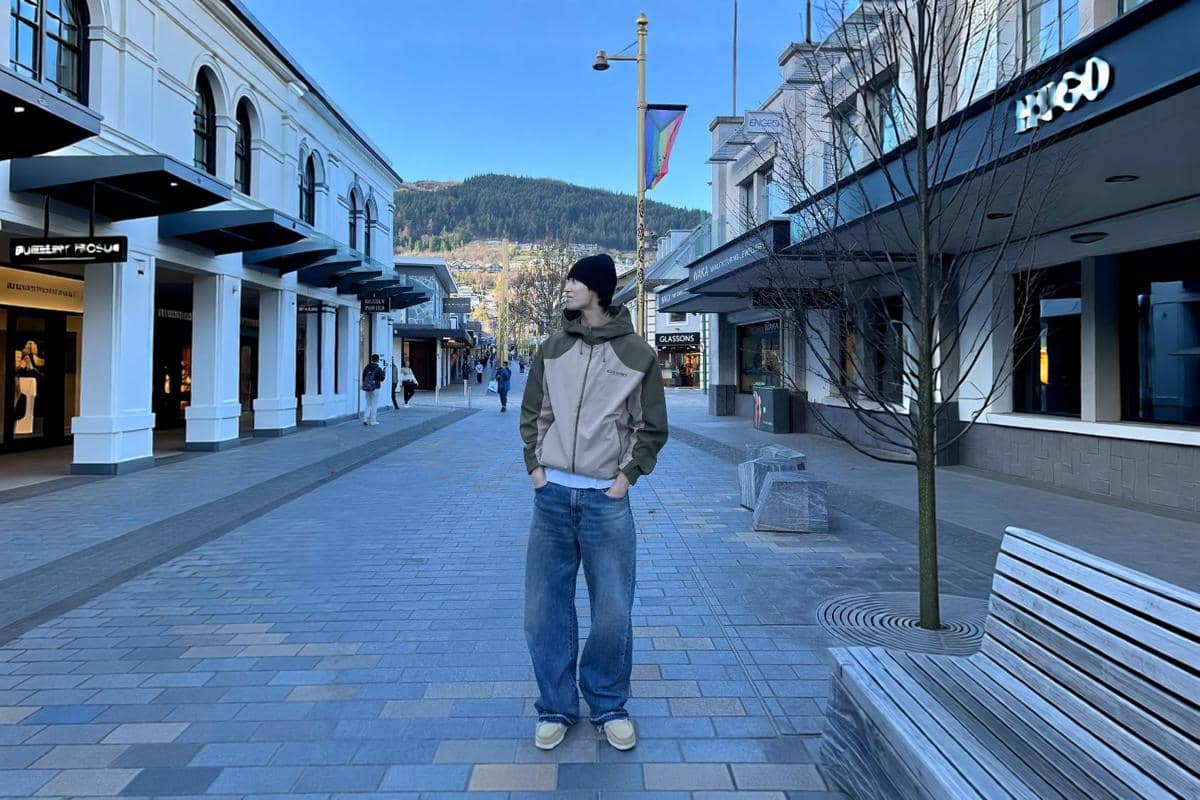 Byeon Woo Seok berdiri di jalanan kota memakai jaket color-block cokelat hijau, celana jeans longgar, dan kupluk hitam
