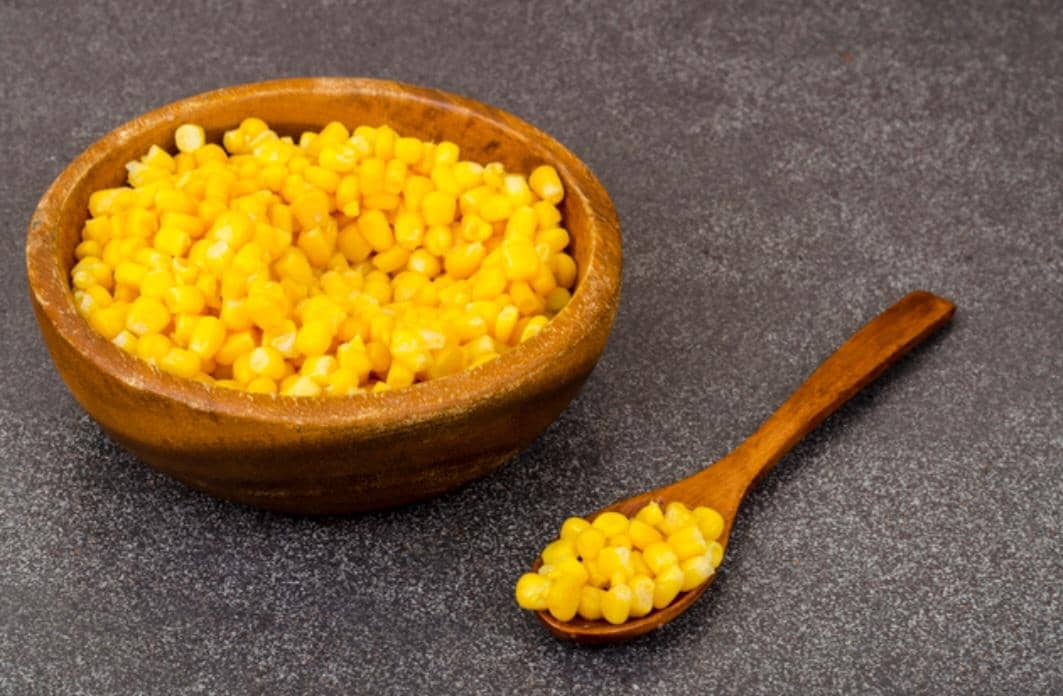 Jagung manis pipil berwarna kuning cerah tersaji dalam mangkuk putih di atas meja kayu dengan latar gelap.