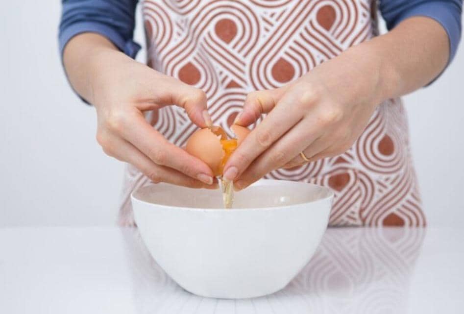 Tangan memecahkan telur ayam di atas mangkuk kaca, dengan kuning dan putih telur jatuh ke dalam wadah.