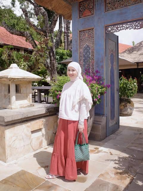 Kesha Ratuliu mengenakan atasan putih dan rok bermotif sambil berpose di pantai dengan latar laut yang cerah.