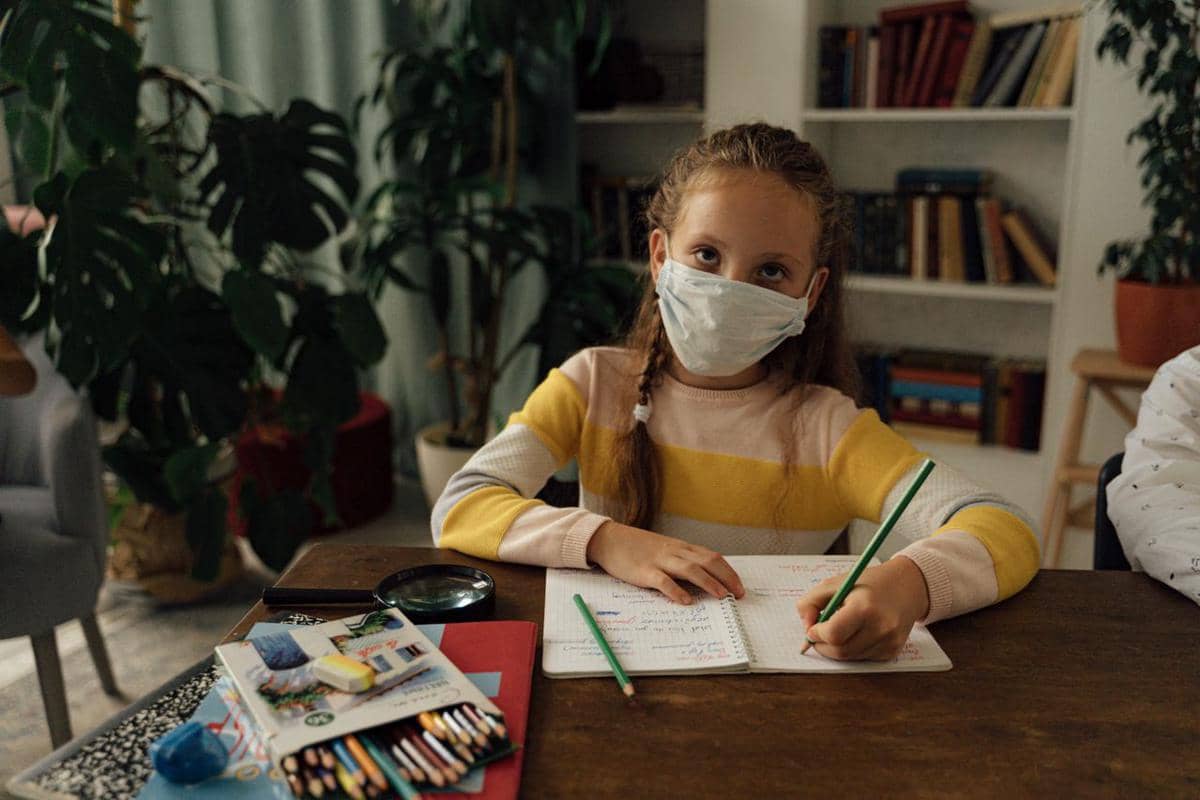 Ilustrasi seorang anak kecil bermasker sedang menulis di buku catatan.