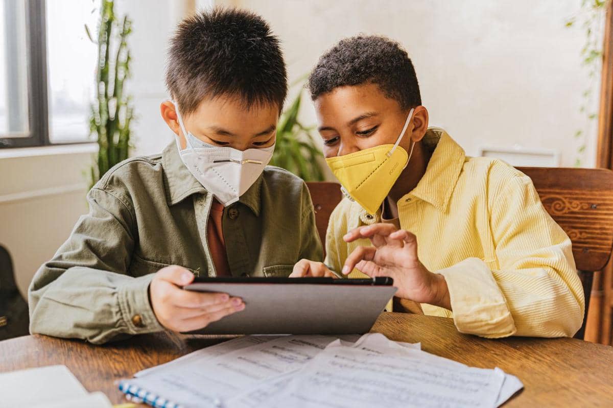 Ilustrasi dua anak laki-laki bermasker menggunakan tablet untuk belajar.
