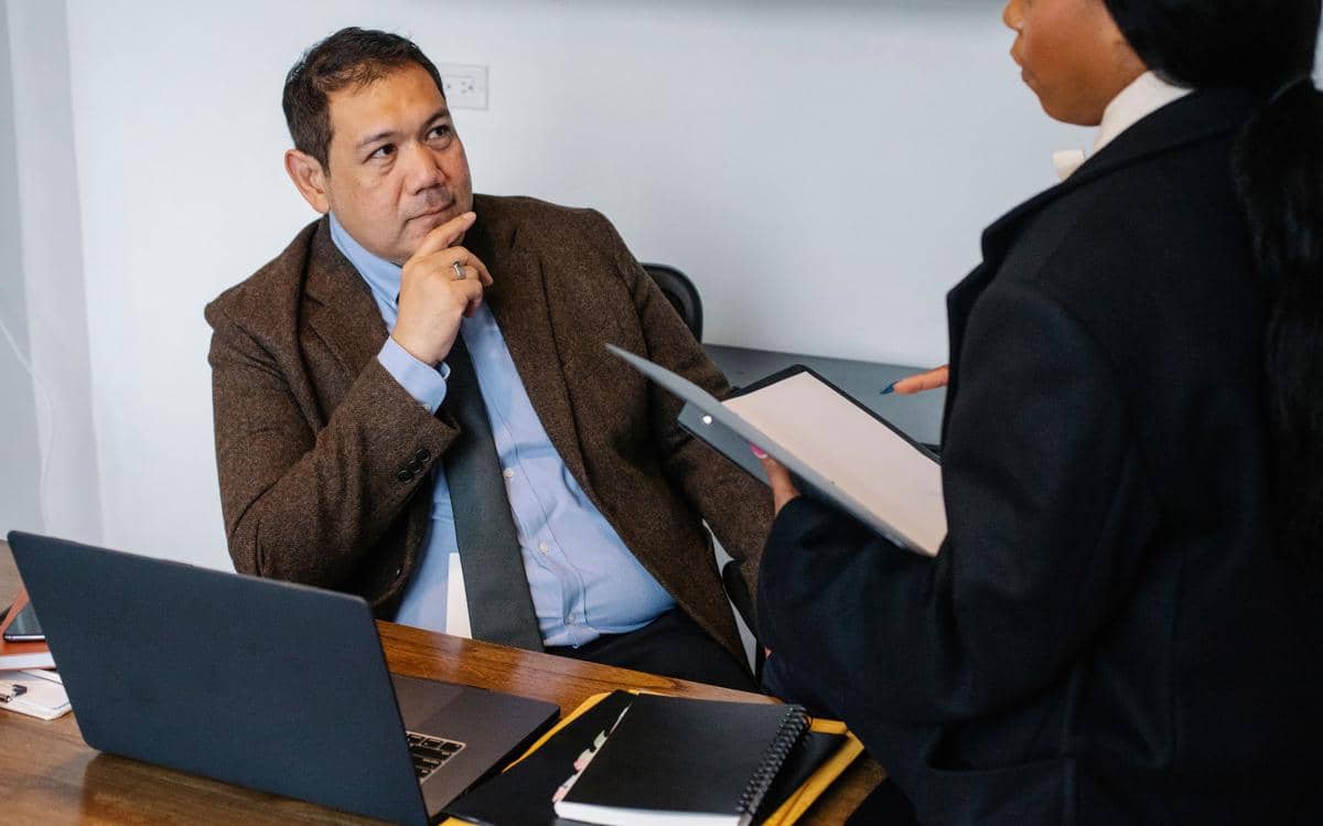 Atasan pria berjas memangku dagu menatap ragu karyawannya yang sedang membawa dokumen di meja kantor