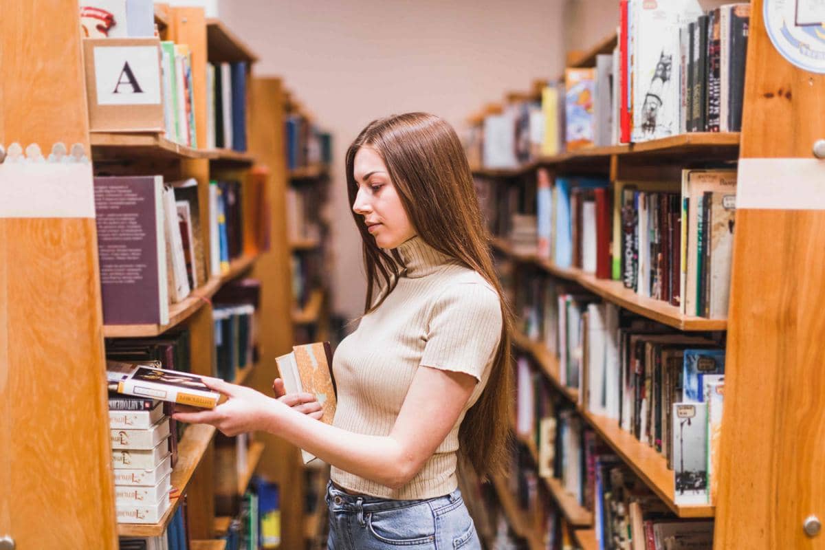 Seorang perempuan belajar sambil membaca buku di perpustakaan, dikelilingi rak buku untuk memilih bacaan sesuai minat.