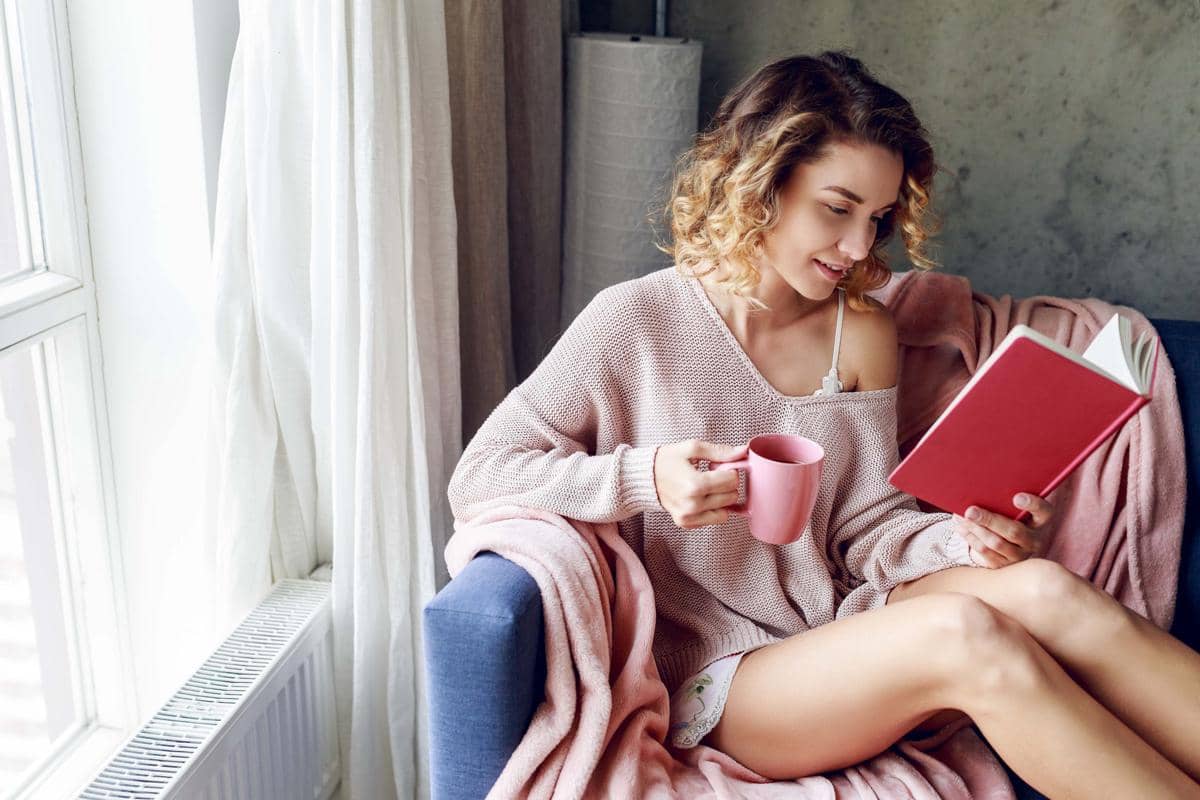 Seorang perempuan tersenyum membaca buku sambil memegang cangkir kopi di rumah yang nyaman pada pagi cerah.