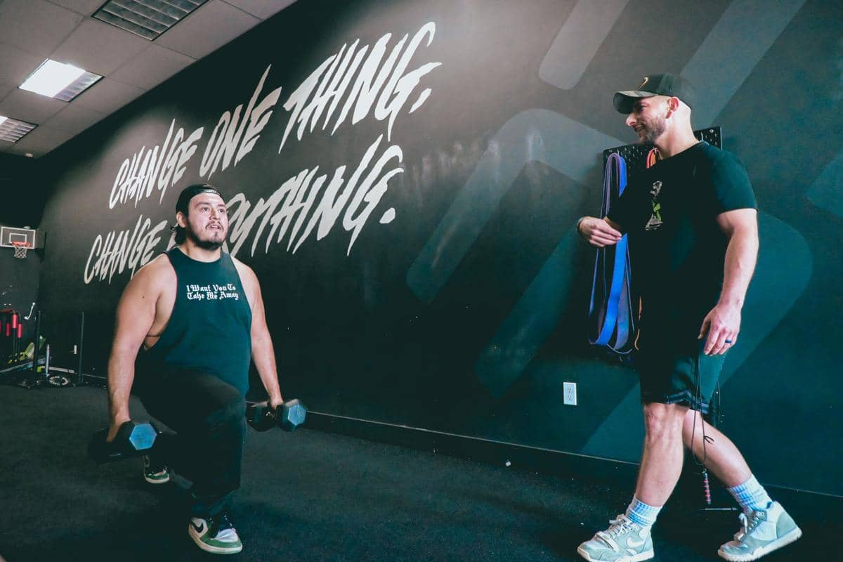 Pria berkaos tanpa lengan memegang dumbbell sambil melakukan gerakan lunge di gym mendampingi pelatih