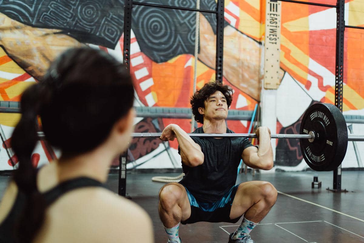 Pria muda berrambut ikal sedang melakukan gerakan barbell squat dengan posisi jongkok di gym
