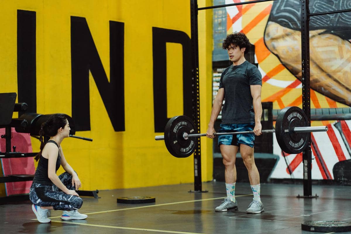 Pria muda berrambut ikal sedang berdiri tegak dalam posisi lockout setelah melakukan deadlift barbell heavy di gym dengan coach watching