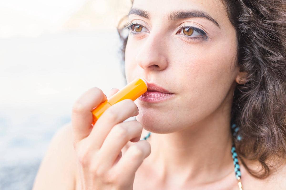 Perempuan muda mengoleskan lip balm pada bibir sebagai ilustrasi penggunaan pelembap bibir ber-SPF harian.