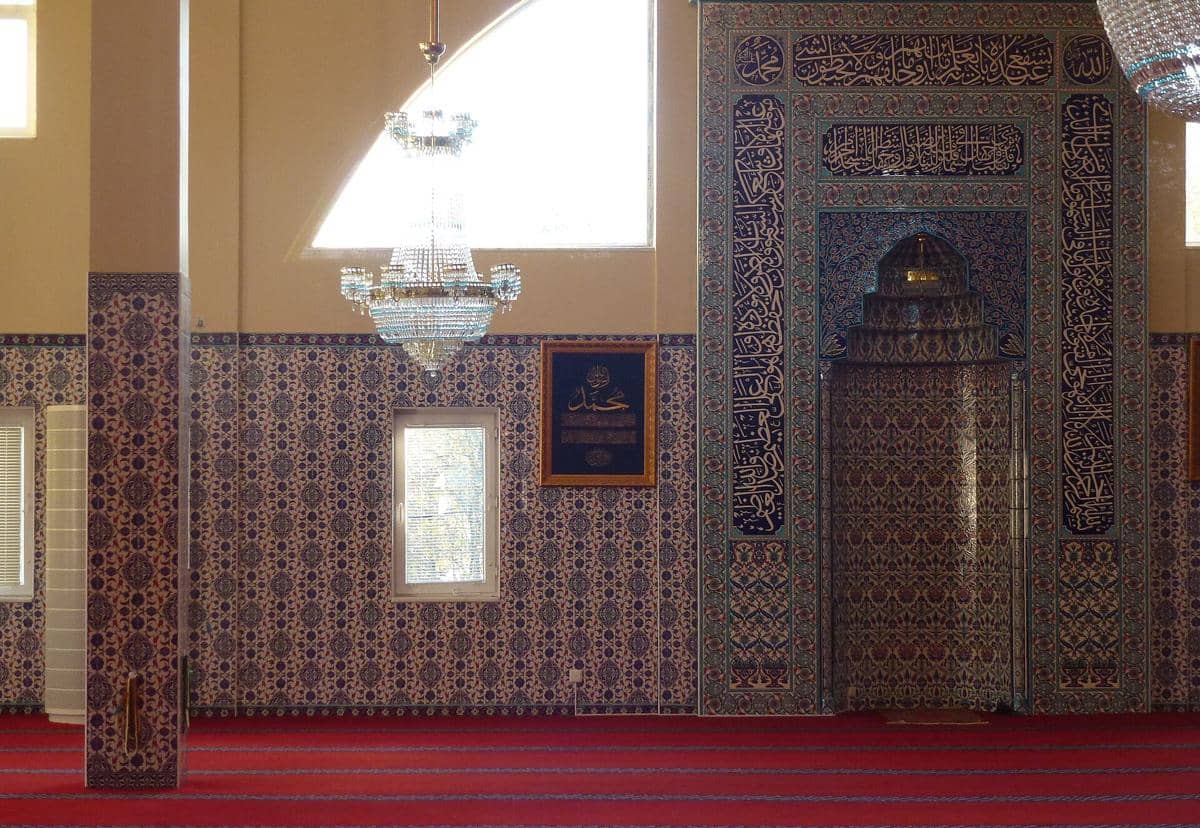 Lampu gantung menghiasi ruang salat di dalam Masjid Fittja, sementara mihrab berornamen berada di dinding depan.