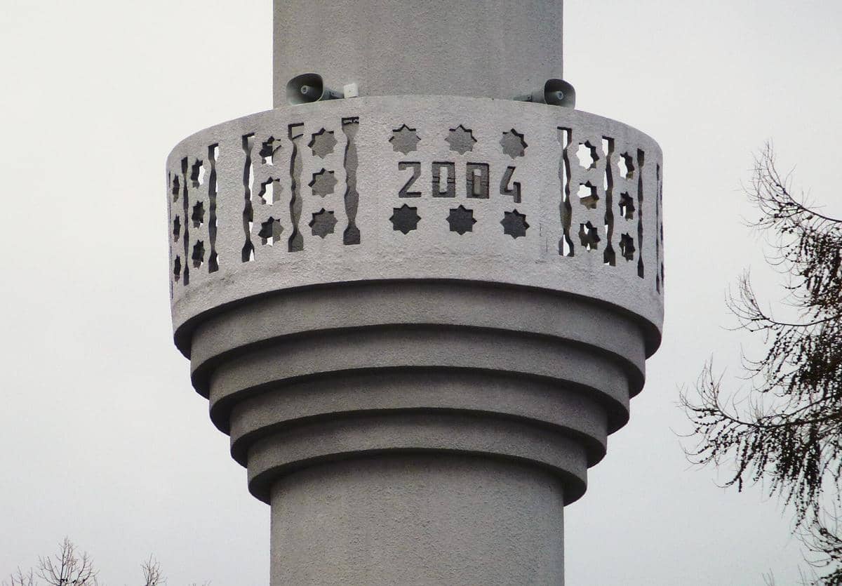 Pengeras suara azan berwarna putih terpasang di menara Masjid Fittja, tampak dari dekat dengan latar langit.