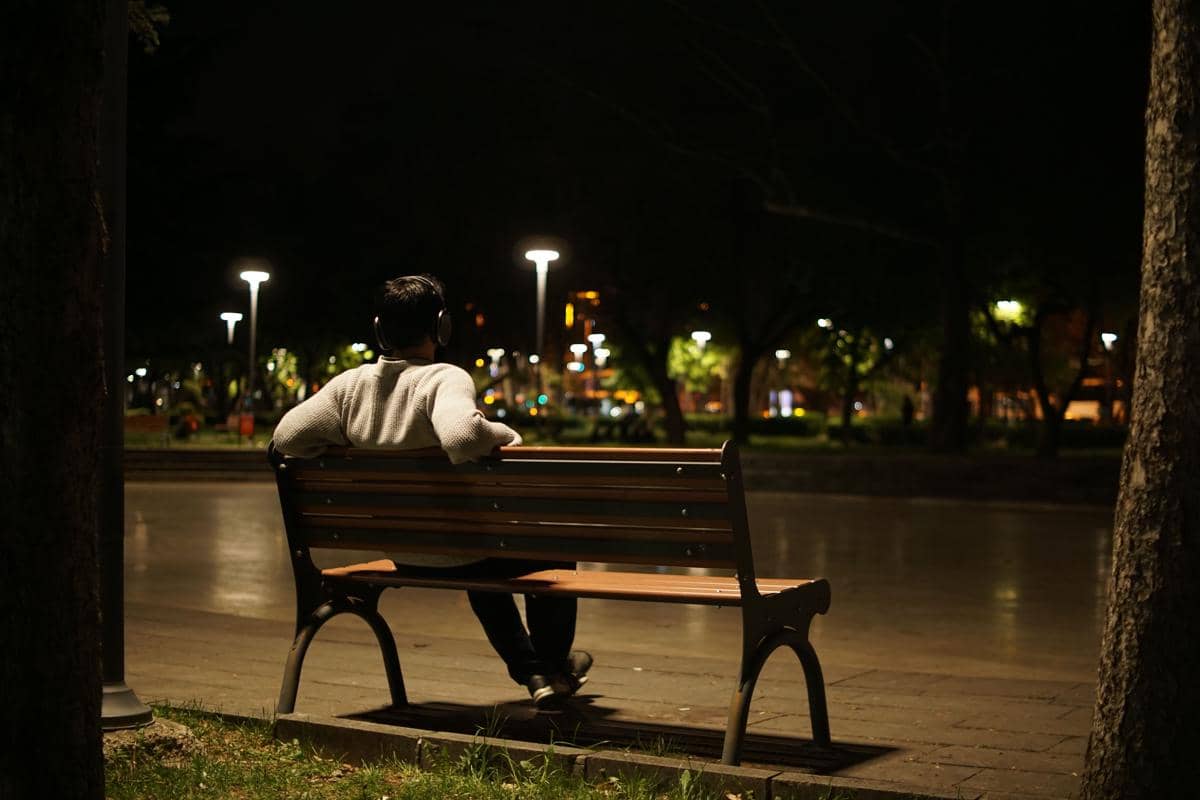 tampak belakang pria duduk sendirian di bangku taman di bawah lampu malam