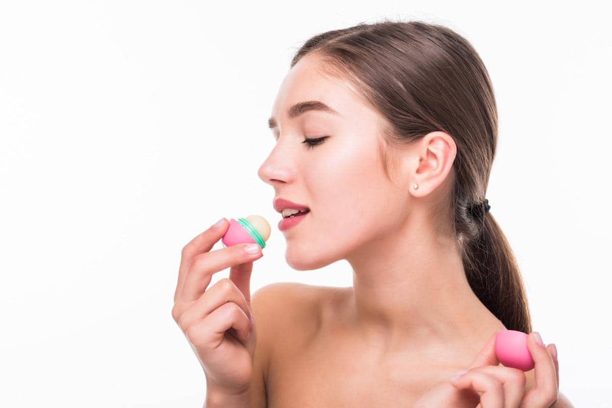 Seorang perempuan muda mengoleskan lip balm pada bibir sambil bercermin dengan latar dinding putih di rumah.