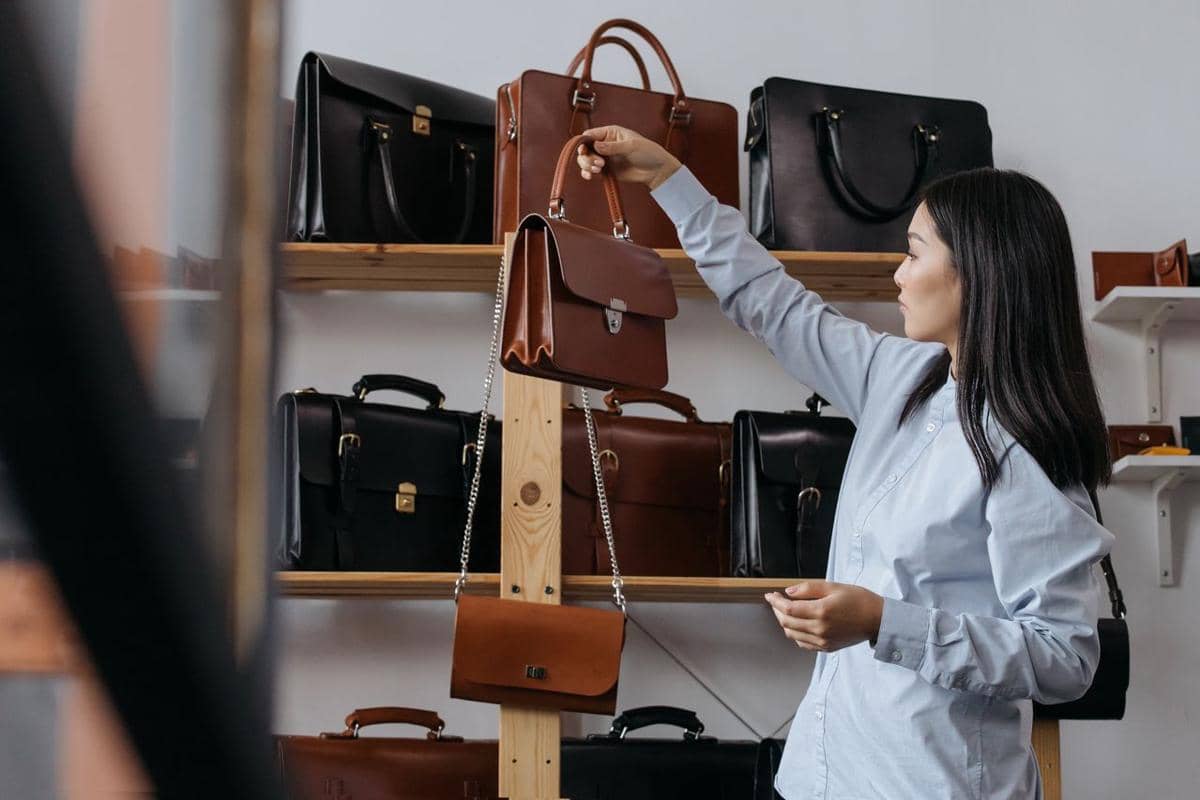 Seorang wanita melihat dan memilih berbagai tas tangan berbahan kulit yang tersusun di rak dalam sebuah toko.